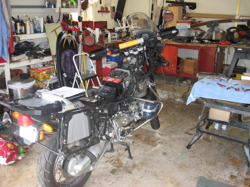 Disassembled BMW R1150GS motorcycle on center stand in a garage, tank removed, engine and wiring exposed.