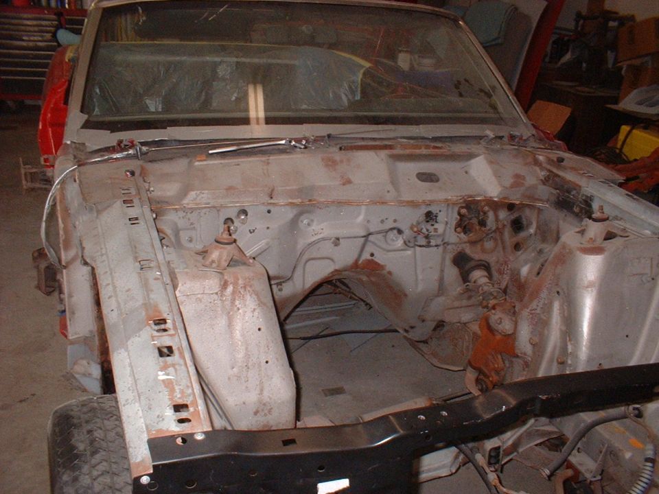 Car engine bay stripped down for restoration. Frame rails and inner fenders are visible, with some rust