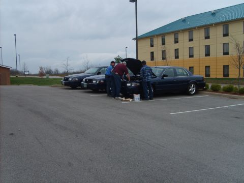 1993 Ford Crown Victoria - Three men attend to the engine of a dark blue 1993 Ford Crown Victoria parked in a lot.