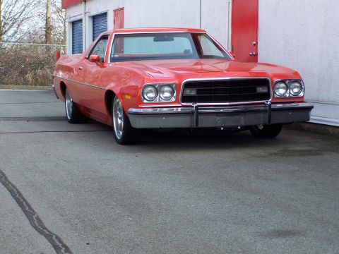 1973 Ford Ranchero - Low front three-quarter shot of an orange-red Ford Ranchero coupe-utility on cracked asphalt beside a white commercial building with red do…