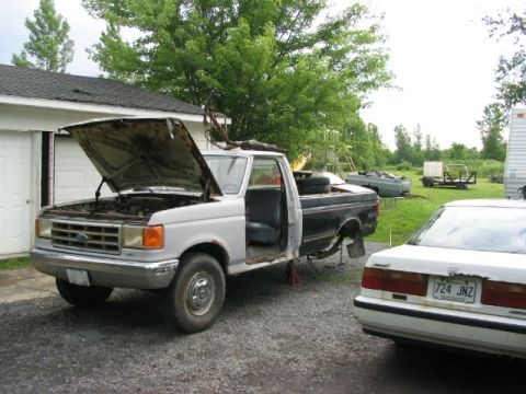 1986 Ford Bronco - A partially disassembled Ford truck sits in a yard next to a white sedan, with a garage in the background.