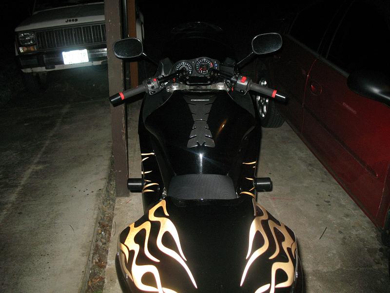 Black motorcycle fuel tank with flame decals, handlebars, and mirrors visible. A Jeep Cherokee is parked in the background
