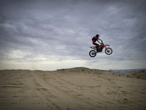 1900 Honda Big Red - A rider on a red dirt bike performs a jump over sand dunes under a cloudy sky.