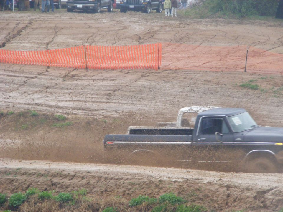 Black 1979 Ford F-250 driving through mud, kicking up spray. Another white truck is behind it