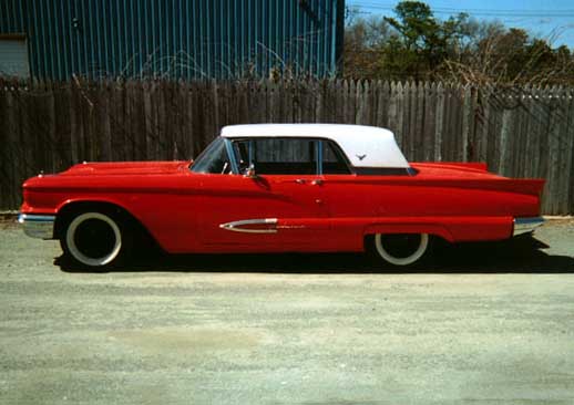 Red late-1950s Ford Thunderbird hardtop with white roof, side profile on gravel by a wooden fence