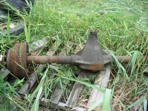 1986 Ford Bronco - A rusty rear axle assembly with a drum brake is seen lying on wooden planks amidst tall grass.