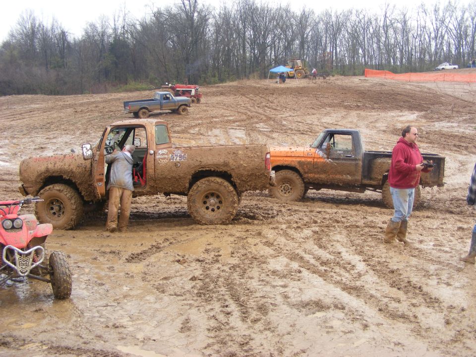 Two muddy pickup trucks, one orange and one brown, stuck in a mud pit with an ATV in the foreground.