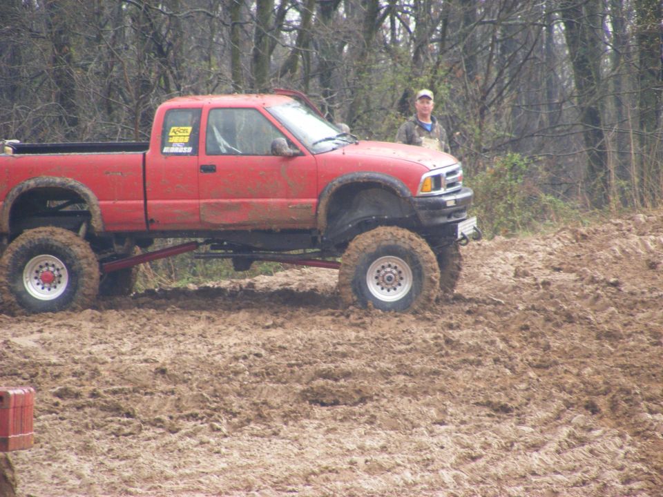 Red lifted pickup truck stuck in mud with a person in the background.