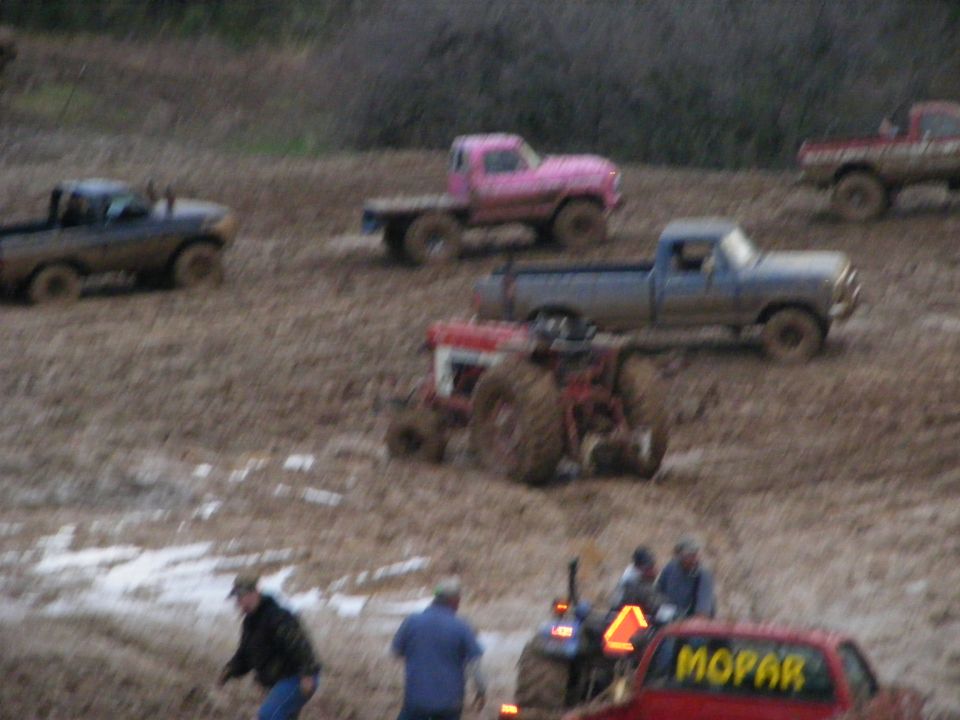 Muddy trucks and a tractor on a brown field. A red truck has "MOPAR" on its rear window.