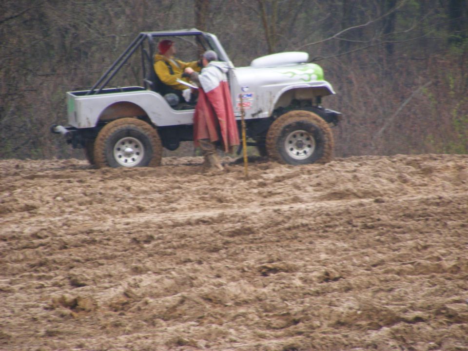 Silver Jeep Wrangler with green flames driving through mud, two people inside, one person outside in a poncho.
