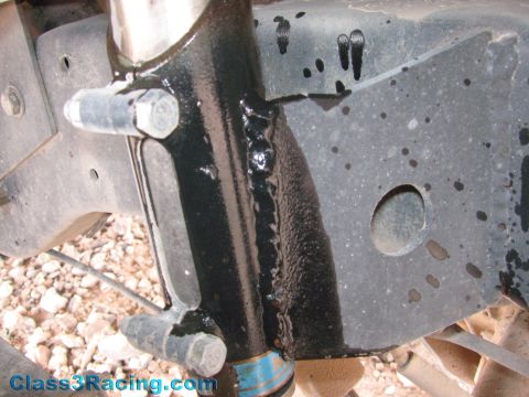 1990 Ford Bronco - Close-up of a freshly wet black-painted vehicle frame and suspension mount with a large bolt, over gravel ground.