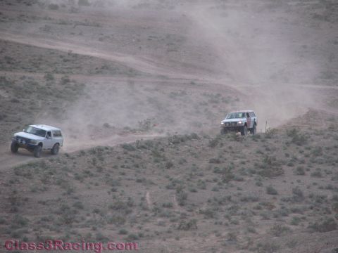 1990 Ford Bronco - Two white off-road SUVs raise dust as they travel along winding desert trails across a barren scrub-covered hillside.