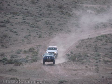 1990 Ford Bronco - Two white off-road SUVs climb a winding dusty desert trail one behind the other across a scrubby hillside.