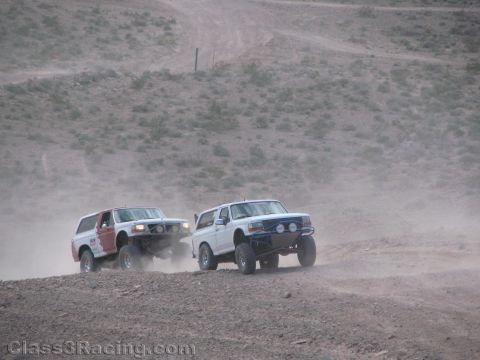 1990 Ford Bronco - Two early-1990s Ford Bronco off-road race trucks, one red and one white, kick up dust climbing a desert dirt trail.