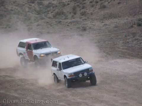 1990 Ford Bronco - pair of white Ford Bronco race trucks throw up dust as they round a curve on a barren desert course.