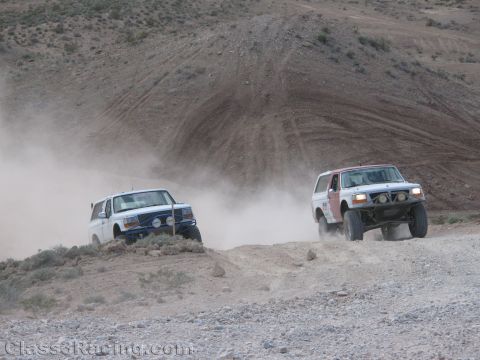 1990 Ford Bronco - Two off-road Bronco race trucks, one blue and one white, charge up a rocky dust-covered desert hillside.