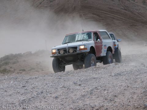 1990 Ford Bronco - white lifted Ford Bronco race truck crawls over loose rocks on a dusty desert hillside, a second truck behind it.