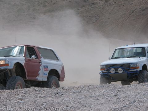 1990 Ford Bronco - Two lifted Ford Bronco race trucks sit side by side on a rocky, dusty desert grade, one showing a Ford Trucks decal.
