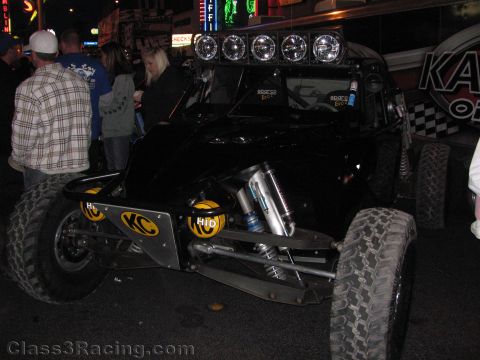 1990 Ford Bronco - black off-road race buggy with a roof light bar and KC and HID lights is displayed on a street at night as people gather.