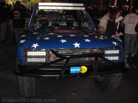 1990 Ford Bronco - front of off-road race truck 760, wrapped in a blue stars-and-stripes hood with LED light bars, sits at a night street event.