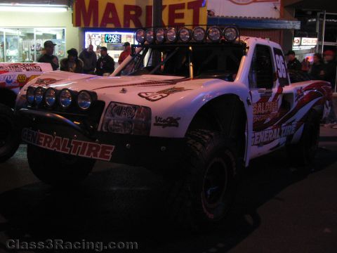 1990 Ford Bronco - white General Tire off-road race truck with a roof light bar sits before a lit market storefront at a night street event.