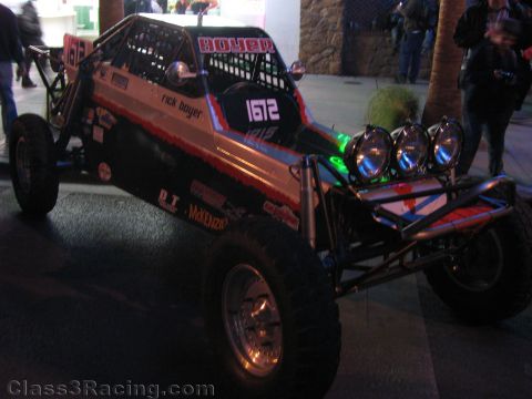1990 Ford Bronco - Off-road race buggy 1612, lettered for Rick Boyer with a row of front light pods, is displayed on a paved area at night.