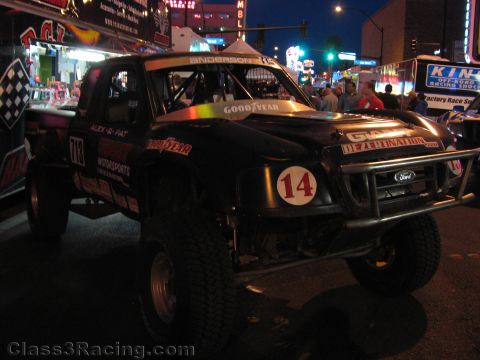 1990 Ford Bronco - Black Ford off-road race truck number 14 with Goodyear graphics sits on a neon-lit street at night with onlookers nearby.