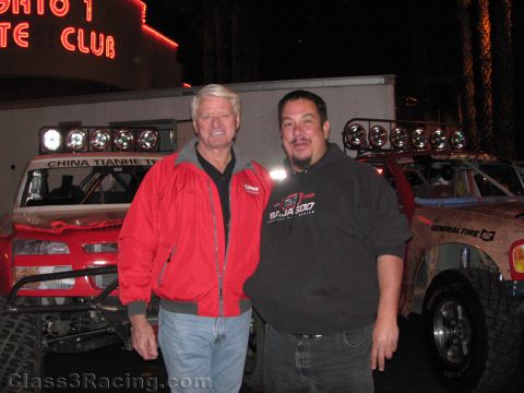 1990 Ford Bronco - Two men, one in a red jacket and one in a Baja 500 sweatshirt, pose in front of an off-road race truck under a neon club sign.