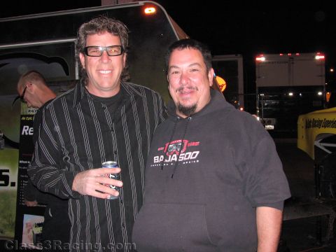 1990 Ford Bronco - Two smiling men pose at a nighttime event near parked trailers, one in a striped shirt holding a drink can and one in a Baja 500 hoodie.
