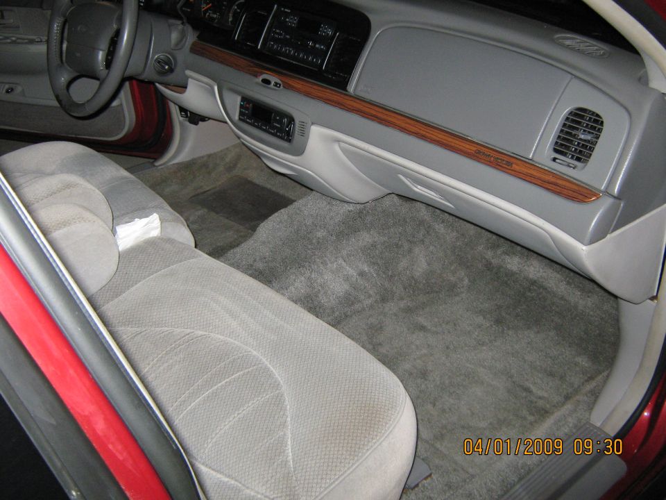 1997 Ford Crown Victoria interior with grey cloth seats, wood trim dashboard, and floor mats. Red exterior visible on the door frame