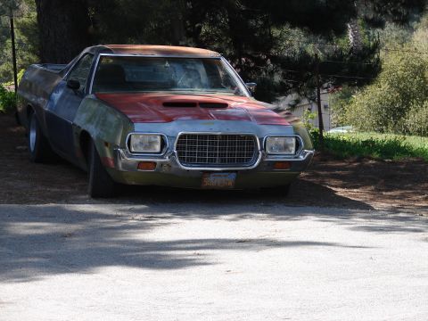 1973 Ford Ranchero - A 1970s Ford Ranchero coupe utility sits on a gravel drive in mismatched primer, faded red and gray body panels with surface rust on the ro…