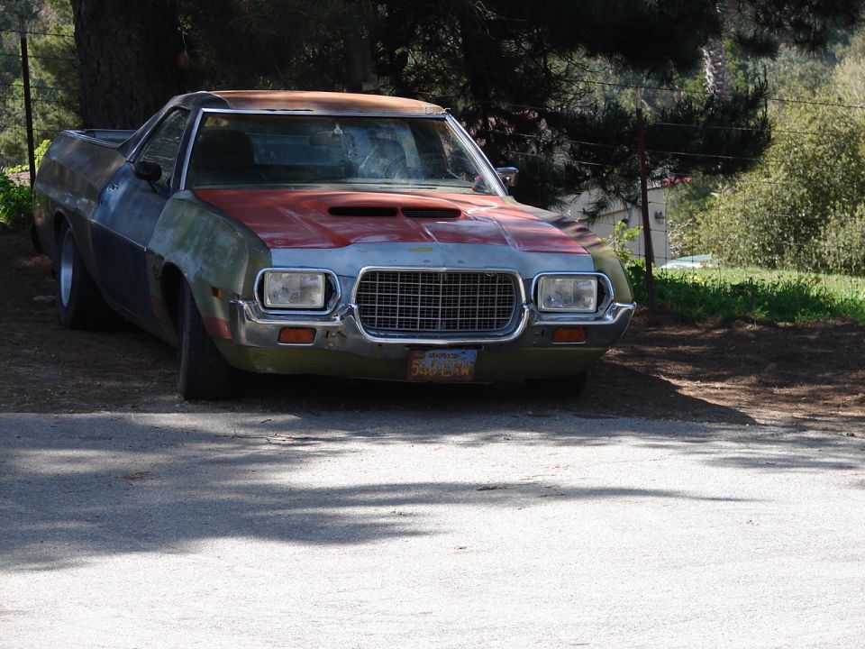 Weathered primer and red 1970s Ford Ranchero parked on a gravel drive, front view