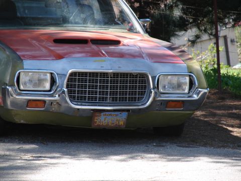 1973 Ford Ranchero - Front-end close-up of an early-1970s Ford in patchwork paint — olive green body, orange hood with twin hood scoops, and gray primer across…