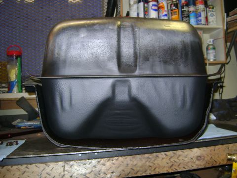 1989 Ford Bronco - A black metal fuel tank is shown from the side, sitting on a workbench with shelves of automotive supplies in the background.