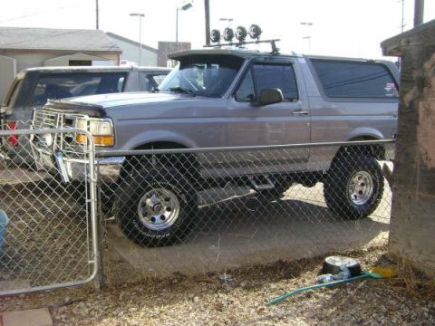 1994 Ford Bronco - A silver Ford Bronco is parked next to another vehicle, partially obscured by a fence.