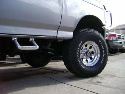 1994 Ford Bronco - A silver 1994 Ford Bronco is shown from the side, highlighting its off-road tires and step bars, with another vehicle visible in the backgr…