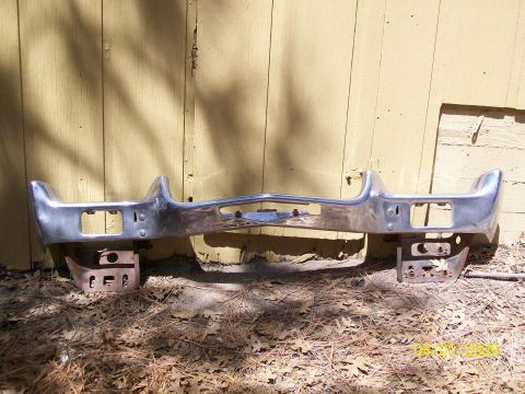 1972 Ford Ranchero - A chrome bumper, likely for a classic car, rests on rusty supports against a weathered wooden wall.