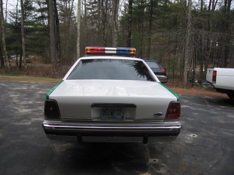 1991 Ford LTD Crown Victoria - Rear view of a white-and-green Ford sedan fitted with a roof lightbar, chrome bumper and a 35LWP72 plate, with a wagon and white truck near…