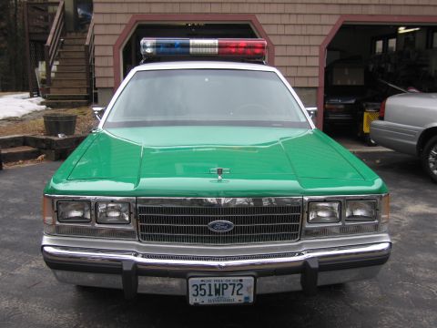 1991 Ford LTD Crown Victoria - Front view of a green-and-white Ford sedan with a roof lightbar, dual headlights and a 351WP72 New Hampshire plate, parked by a garage.