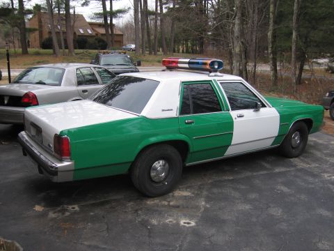 1991 Ford LTD Crown Victoria - Rear driver-side view of a green-and-white Ford patrol-style sedan with a roof lightbar parked on a driveway near other cars and a tow truc…
