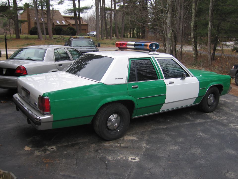 Rear driver-side view of green-and-white Ford sedan with roof lightbar