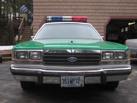 1991 Ford LTD Crown Victoria - Close front view of a green-hooded Ford sedan showing the chrome grille, Ford oval, light bar, and 351WP72 plate.