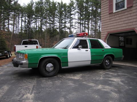 1991 Ford LTD Crown Victoria - green and white full-size Ford sedan with roof light bar parked in profile beside a shingled house and pickup truck.