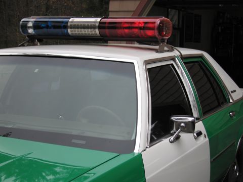 1991 Ford LTD Crown Victoria - red, clear and blue police light bar mounted on the roof of a green and white sedan, viewed close over the windshield.