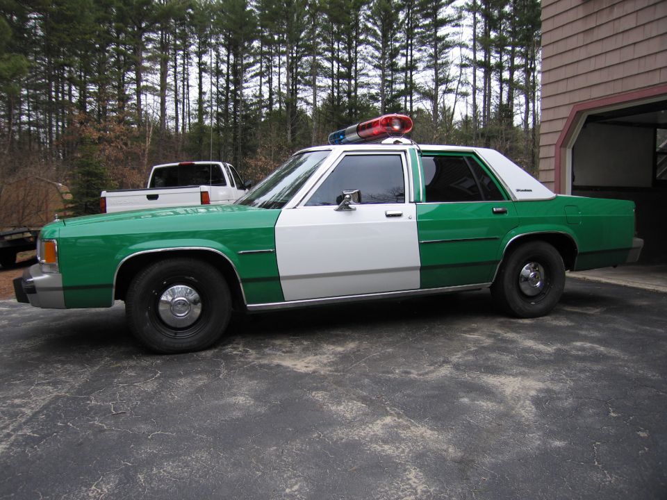 Green and white Ford police sedan, side profile near a house