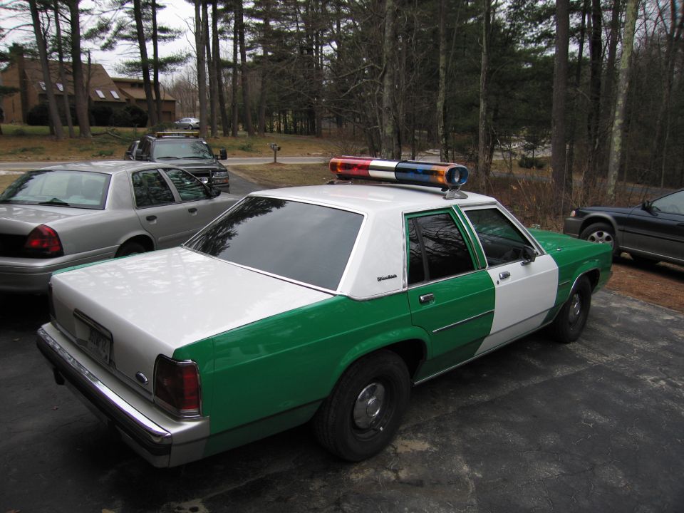 Green and white Ford police sedan, high rear-quarter view in driveway