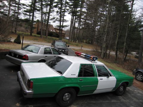 1991 Ford LTD Crown Victoria - green and white full-size Ford sedan with roof light bar viewed from above at the rear, parked near other cars on a driveway.