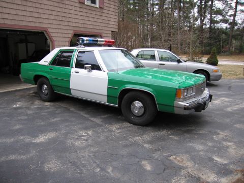 1991 Ford LTD Crown Victoria - green-and-white Ford police-style sedan with roof light bar parked in a driveway beside a shingled house and a silver car.