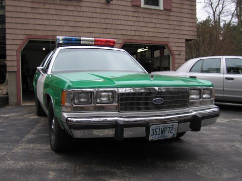 1991 Ford LTD Crown Victoria - Front view of a green-and-white Ford police sedan with roof light bar and New Hampshire plate 351WP72, parked outside a garage.