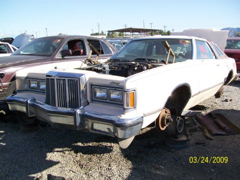 1972 Ford Ranchero - A white 1977 Mercury Cougar sedan sits in a junkyard with its hood open and missing wheels.